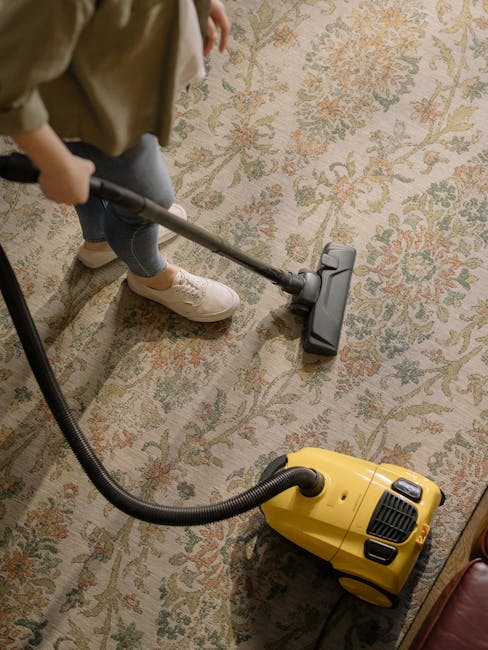 A person using a vacuum cleaner on a large, ornate area rug with a floral pattern in a well-lit living room. The individual is dressed in casual clothing and white sneakers, pushing the vacuum across 