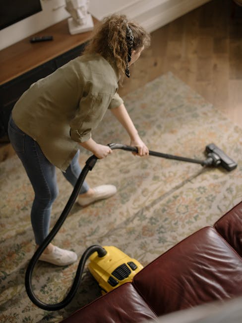 A person using a vacuum cleaner on a large, ornate area rug with a floral pattern in a well-lit living room. The individual is dressed in casual clothing and white sneakers, pushing the vacuum across 