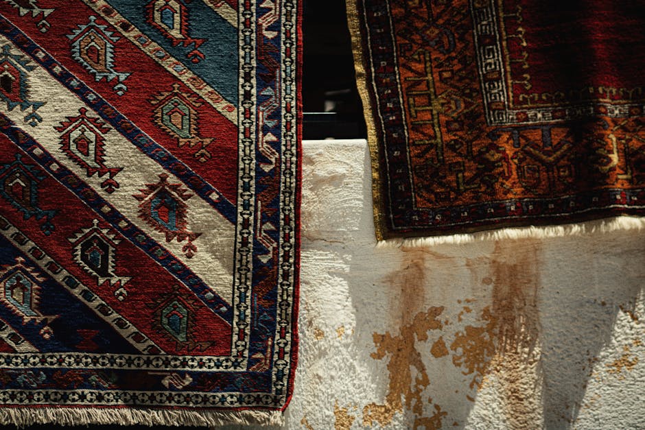 A green and beige patterned Persian-style rug with intricate floral and geometric designs, positioned vertically on an outdoor concrete surface, in front of a weathered brick and plaster wall with vis