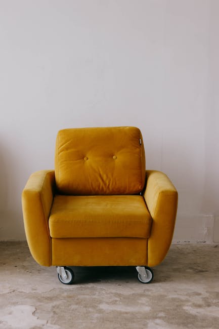 A mustard yellow armchair with a plush, tufted backrest and wide armrests, placed on a smooth, light grey concrete floor against a plain white wall. The armchair features caster wheels underneath, all